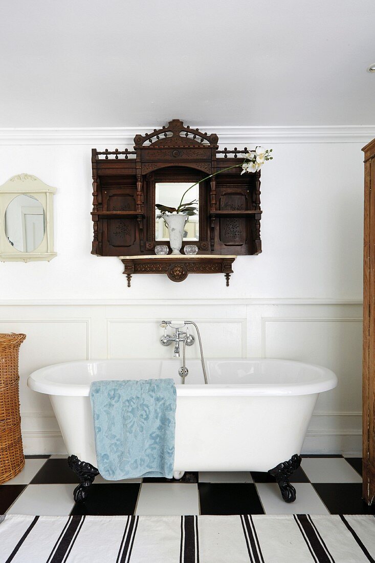 Antique wall-mounted cabinet made from dark wood above clawfoot bathtub and black and white rug on chequered tiled floor