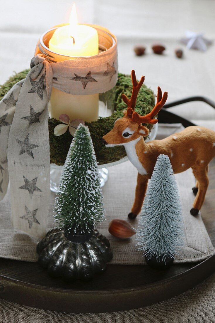 Christmas arrangement of miniature Christmas trees, deer ornament and lit candle in glass inside moss wreath