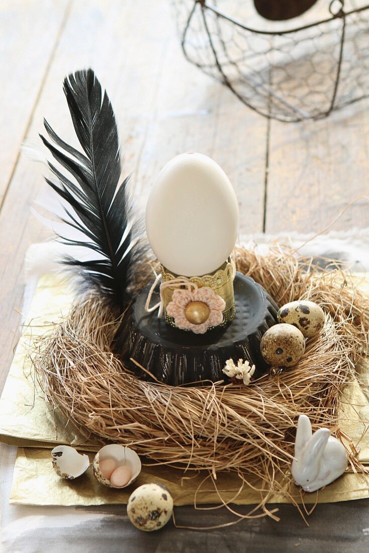 Vintage-style Easter arrangement of duck's egg on upturned cake tin in straw nest with quail's eggs and black feather