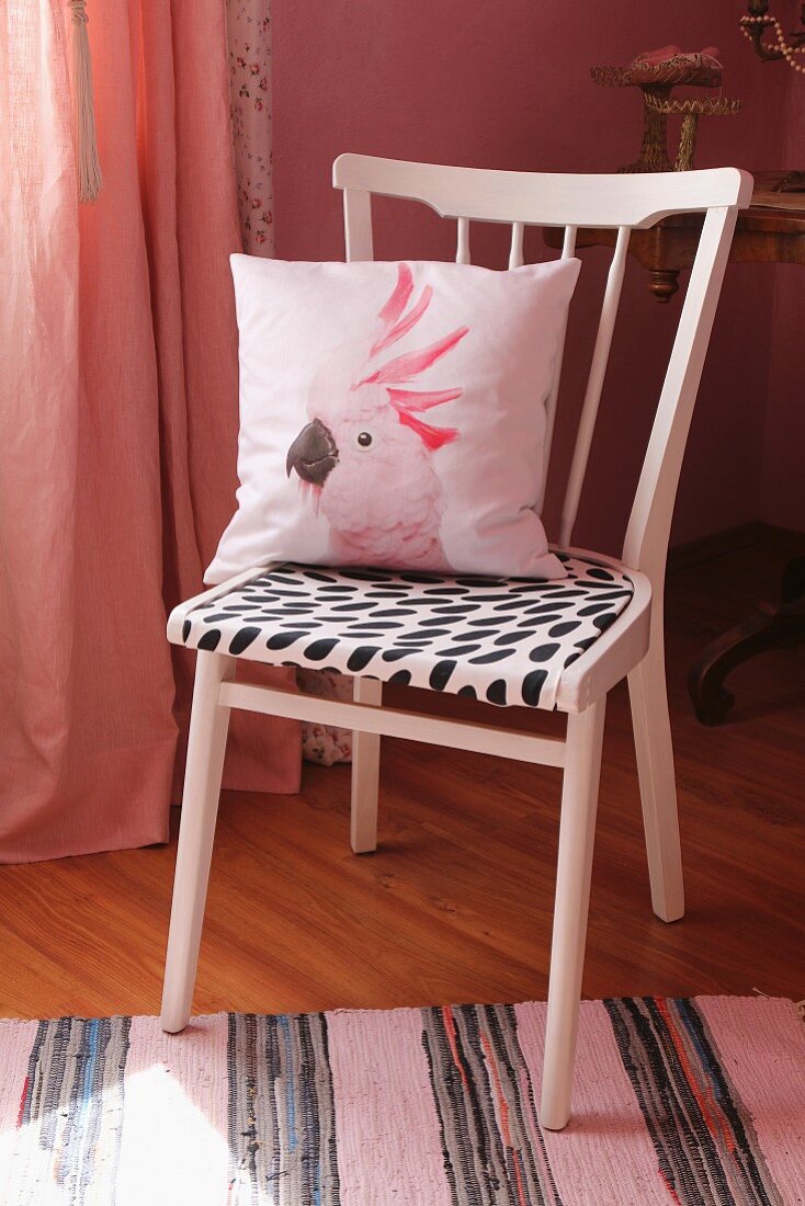 Upcycling: Rosa lackierter Küchenstuhl mit schwarz-weißem Sitzpolster und Kissen mit Kakadu-Abbildung