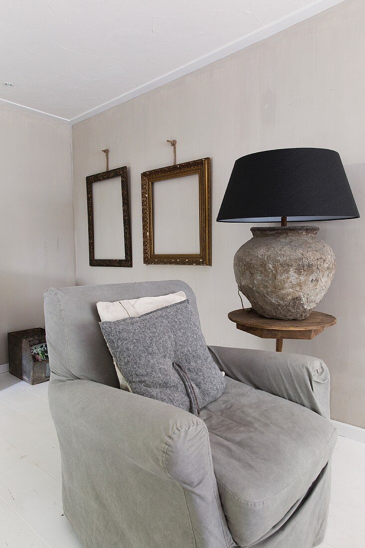 Grey armchair next to rustic table lamp on side table in front of empty picture frames on wall painted pale grey