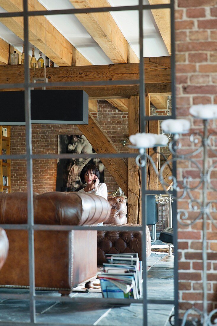 Blick durch Metallgitter auf Frau in Loungebereich einer Loft-Wohnung mit Ziegelwänden und Holzbalkendecke