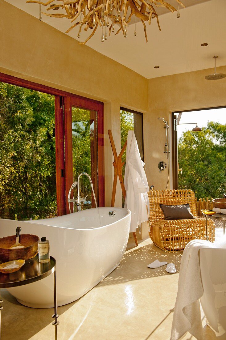 Beig getöntes wohnliches Bad mit freistehender Badewanne und Korbsessel vor offen stehenden Terrassentüren