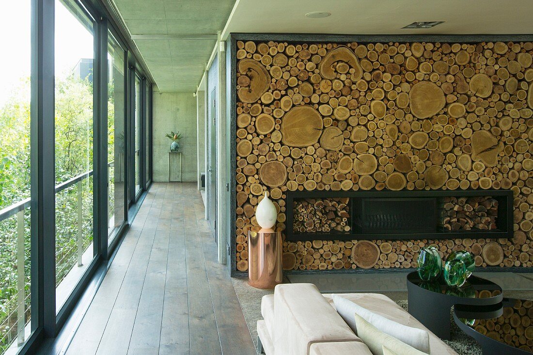 Cylindrical copper table in front of fireplace in wall decorated with slices of tree trunks and next to hallway with glass wall
