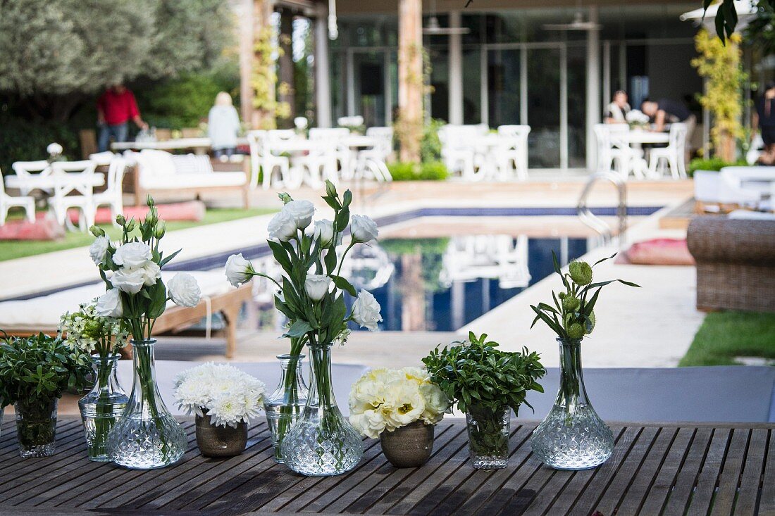 White flowers in crystal vases on wooden table in front of pool and several seating areas