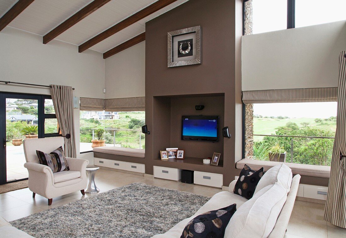 Grosszügiger Wohnraum mit weissen Polstermöbeln um Flokati, Wandnische mit Fernseher zwischen Fenstern mit Sitzpolsterauflagen und eingebauten Schubladen