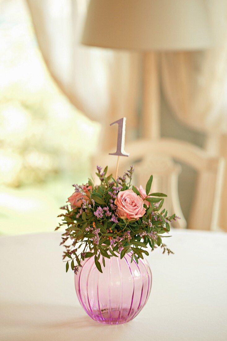 Kugelförmige Glasvase mit Rosengesteck und eingesteckter Tischnummer