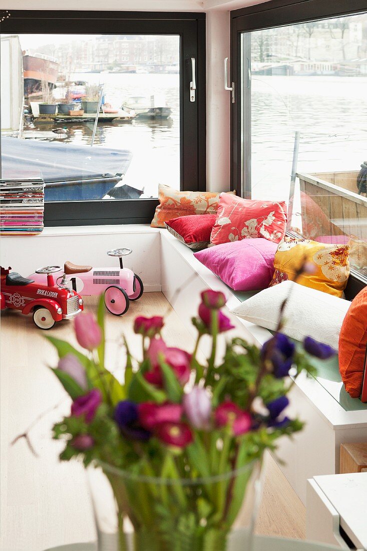 Sitzbank mit bunten Kissen vor Fensterfront in einem Hausboot, Blick aufs Wasser