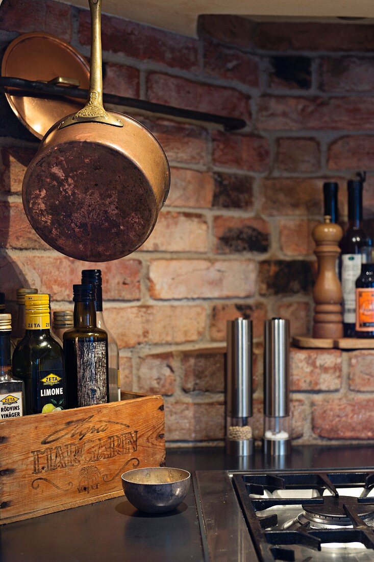 Kupfertöpfe, moderne Gewürzmühlen und Vintage Holzkiste auf Küchenarbeitsplatte hinter Gasherd, rustikale Ziegelwand im Hintergrund