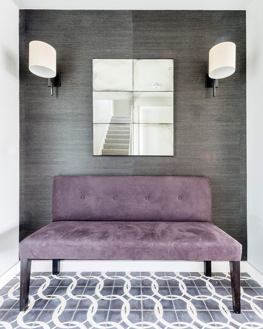 Purple suede couch below mirror flanked by sconce lamps with white lampshades on wall covered in grey wallpaper