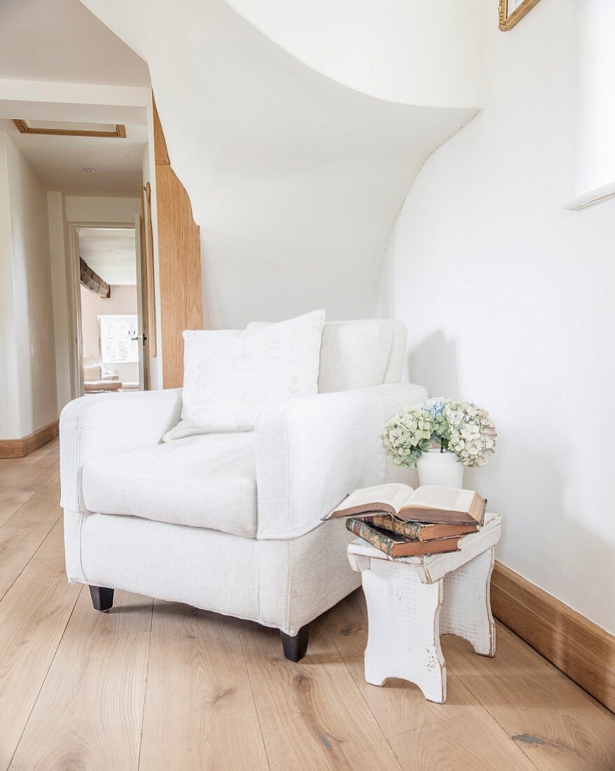 Polstersessel mit weißem Bezug und rustikaler Holzschemel unter Treppenlauf im Hausflur eines Landhauses