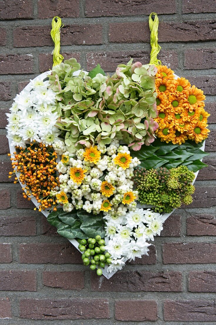Selbst gebasteltes Blumengesteck aus Chrysanthemen, Hortensien und Beeren in Herzform an Ziegelwand aufgehängt