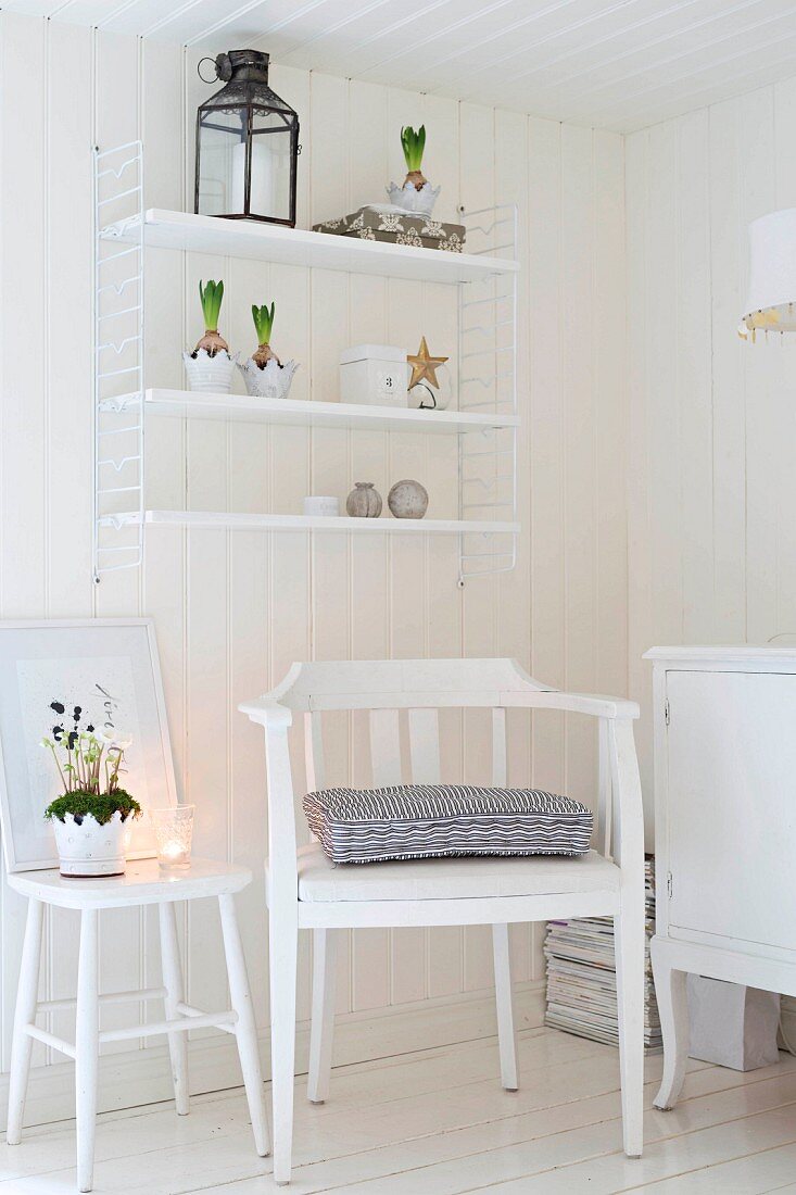 Weiß lackierter Armlehnstuhl und romantisch dekorierter Hocker vor Holzwand mit aufgehängtem String Regal in ländlichem Ambiente