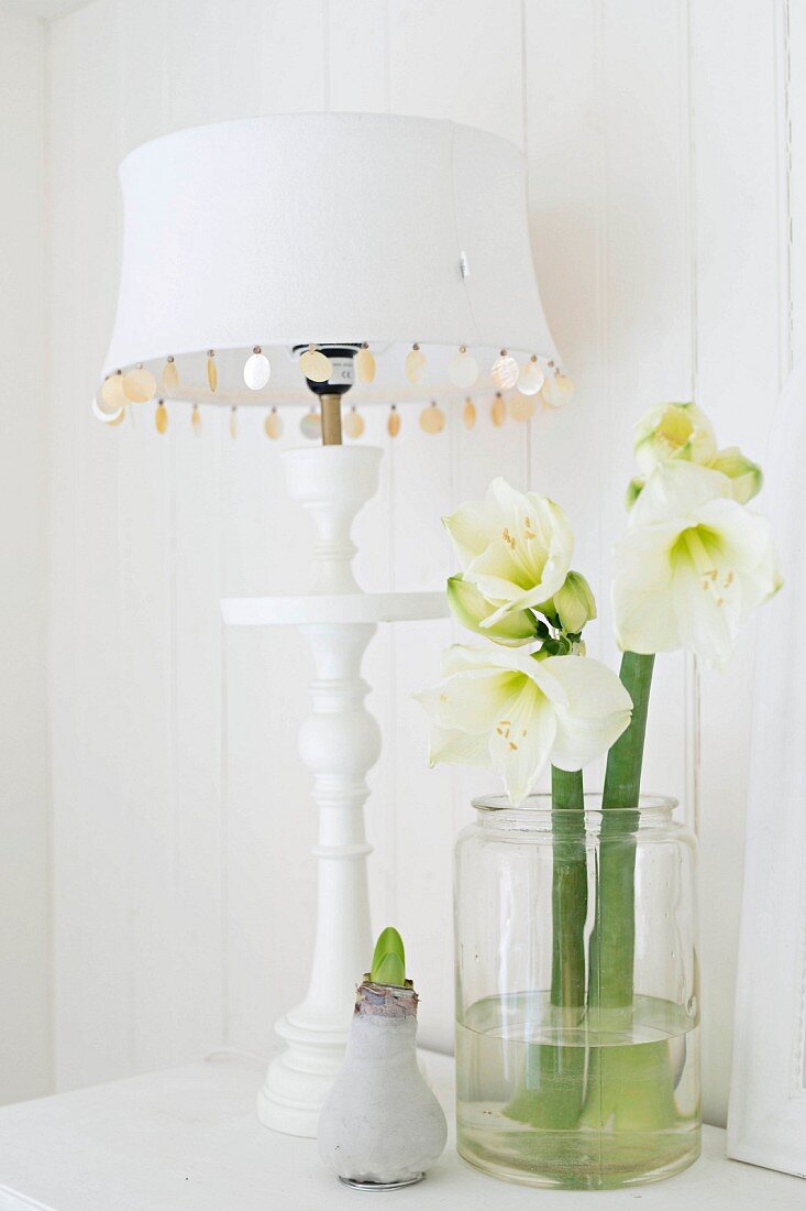 Table lamp with white lampshade and turned base next to glass jar of white amaryllis