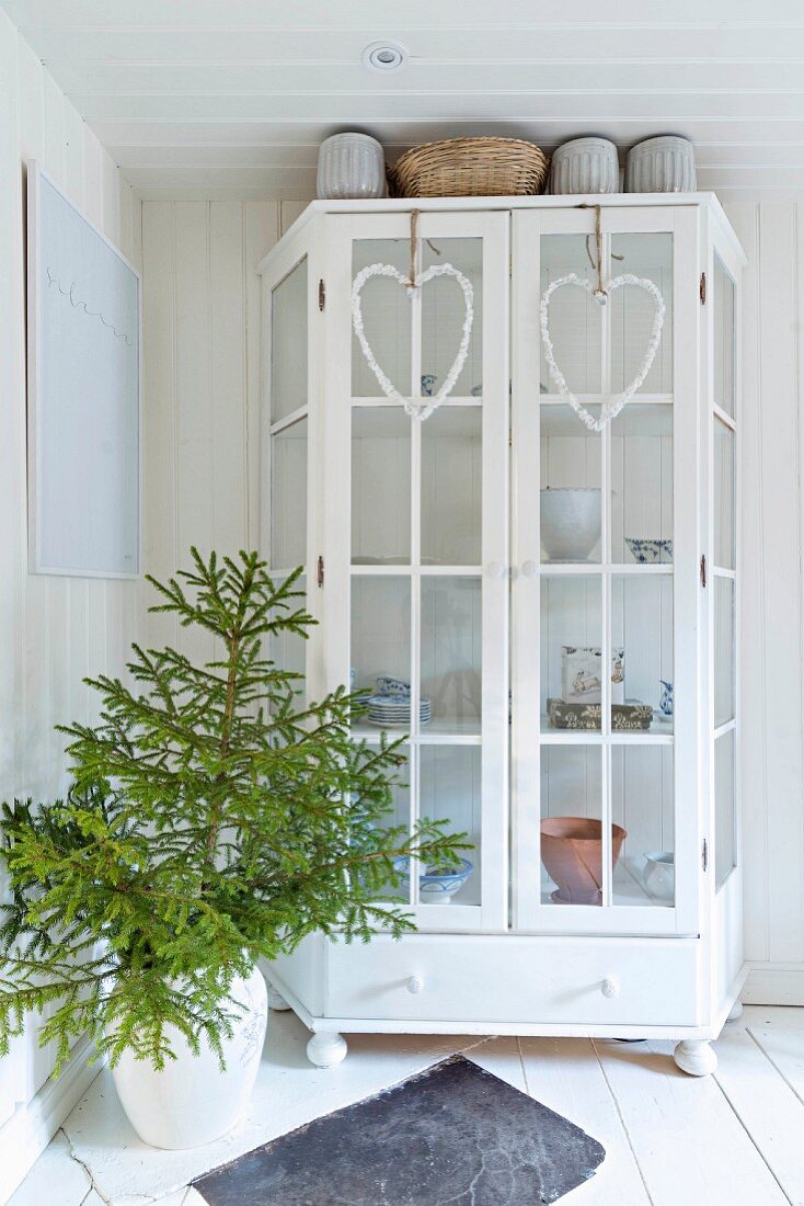 Heart-shaped wreaths hung on doors of white-painted display case next to fir branches in floor vase