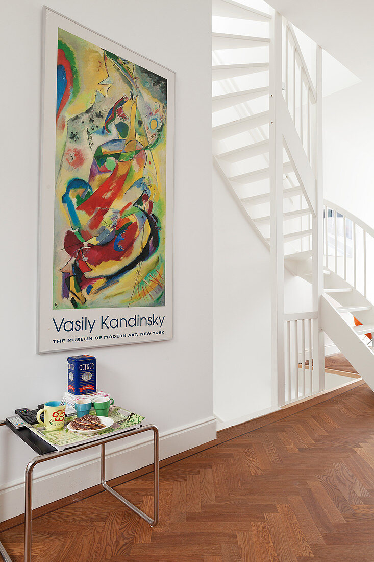 Classic side table below poster on wall of hallway with white, winding staircase to one side