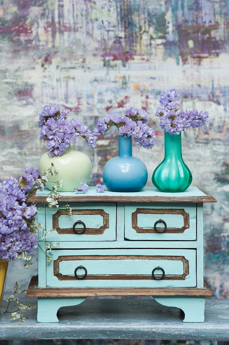 Drei farbige Glasvasen mit lila Trockenblumen auf Vintage Kommode vor malerischem Hintergrund