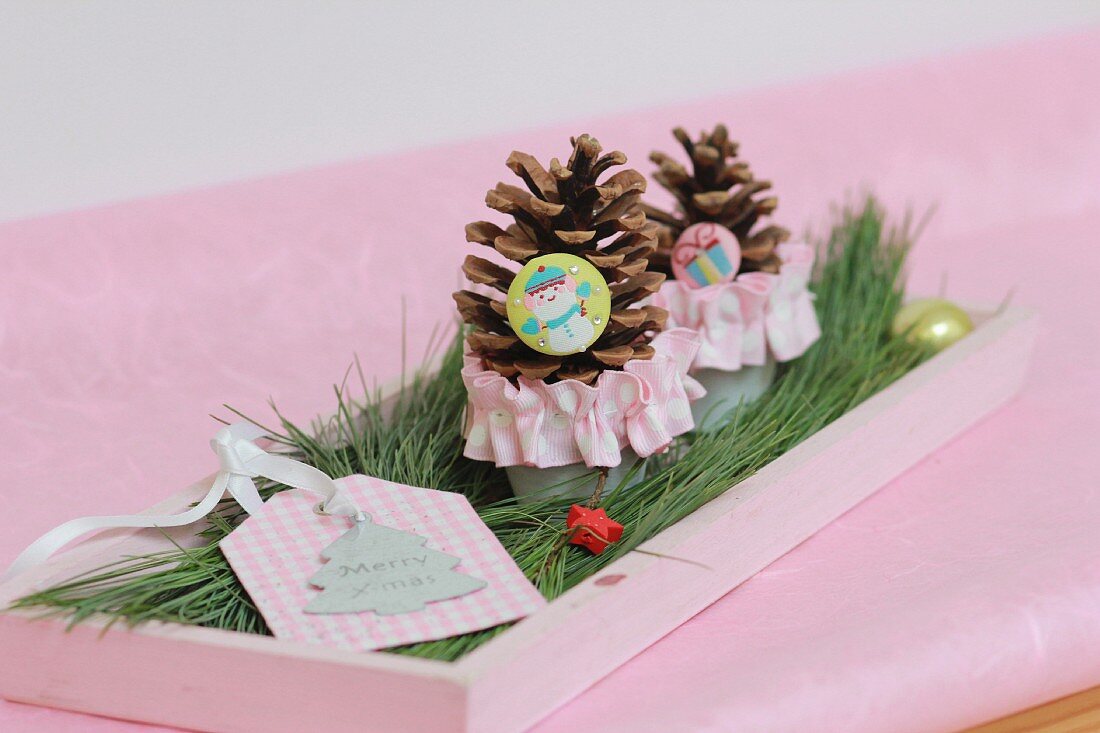 Festive table arrangement of gift tag and pine cones on pink tray