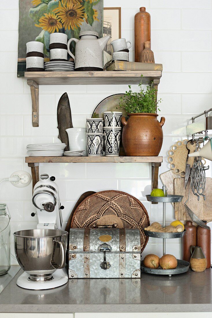 Mixer and vintage bread bin on stone worksurface below crockery on rustic wooden bracket shelves on white wall tiles