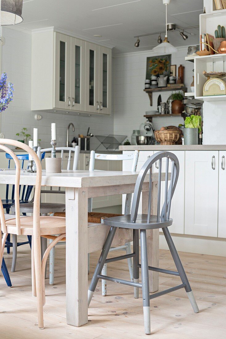 Various chairs around white-painted dining table in fitted kitchen with white-painted, country-house-style kitchen cabinets
