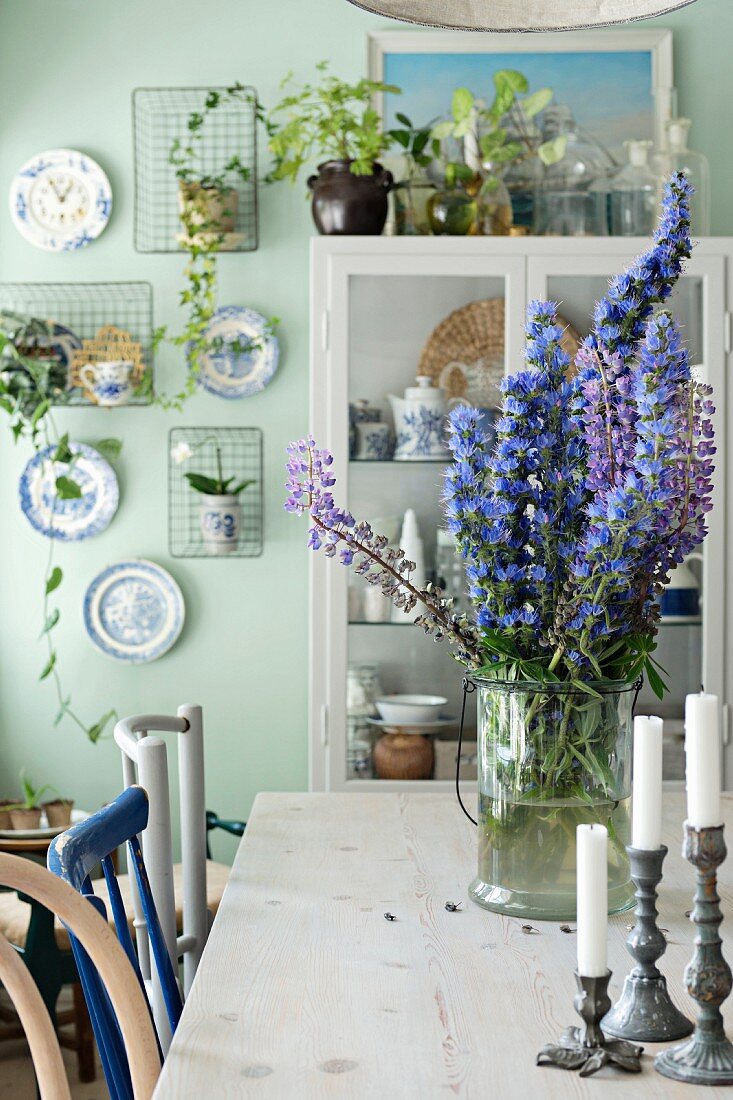 Kerzenhalter und Glasgefäss mit blauen Blumen auf Holztisch, im Hintergrund Vitrine vor hellgrüner Wand und dekorative Wandteller