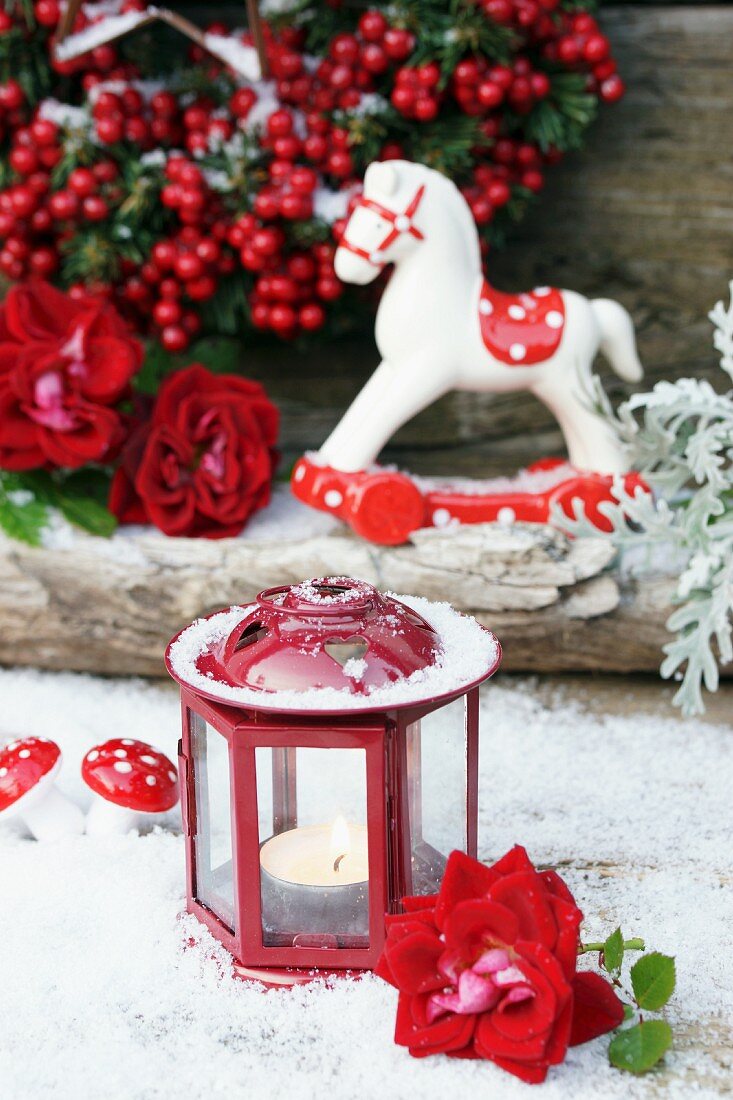 Kleine Windlaterne mit Rosenblüte im Kunstschnee, dahinter ein Deko-Schaukelpferdchen und Winterbeeren