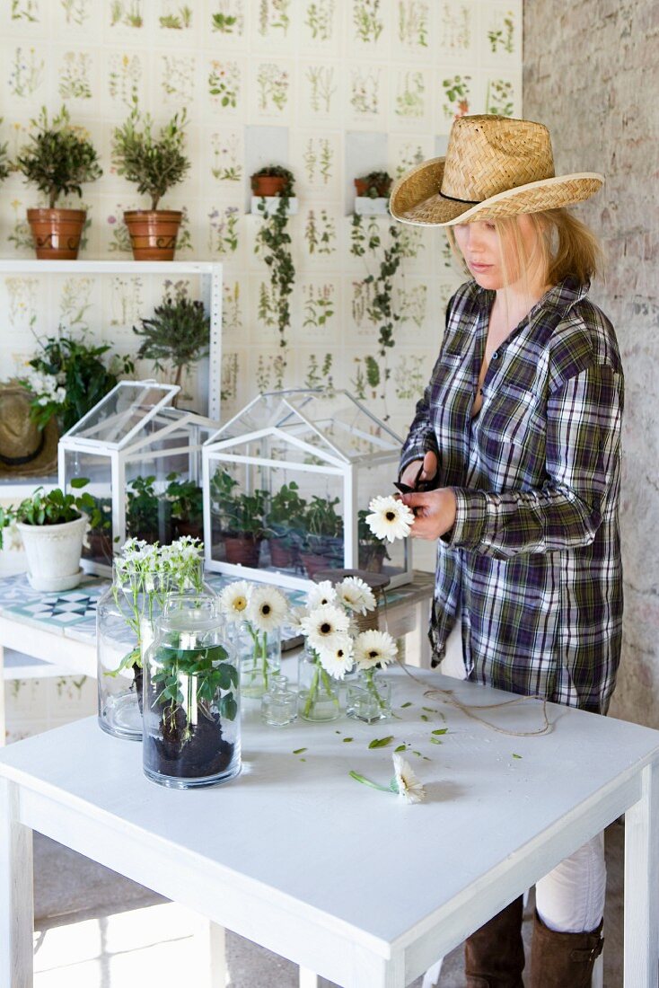 Woman wearing straw hat arranging white gerbera daisies in small glass vases