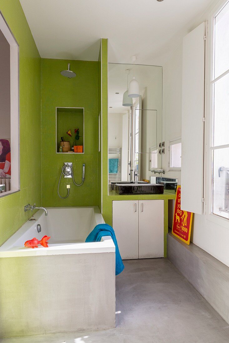 Badewanne, neben Trennwand eingebauter Waschtisch im Badezimmer mit Betonboden und teilweise grünen Wandfliesen