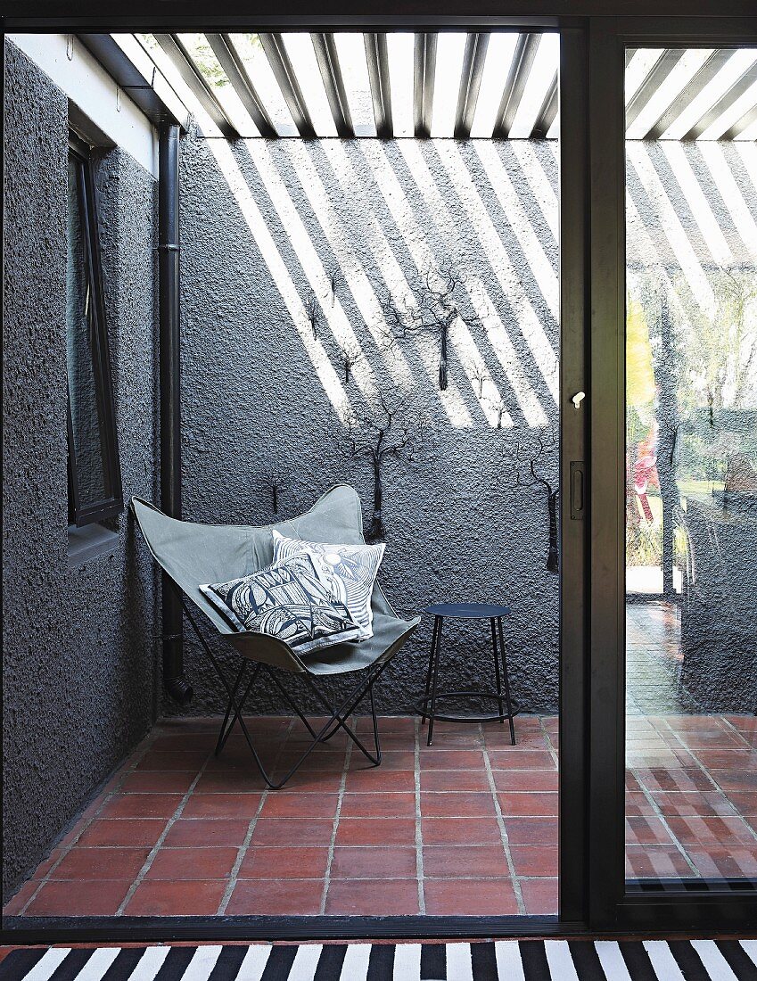 Blick durch Terrassentür auf Butterfly Sessel vor grauer Wand, oberhalb Pergola mit Lamellen Abdeckung