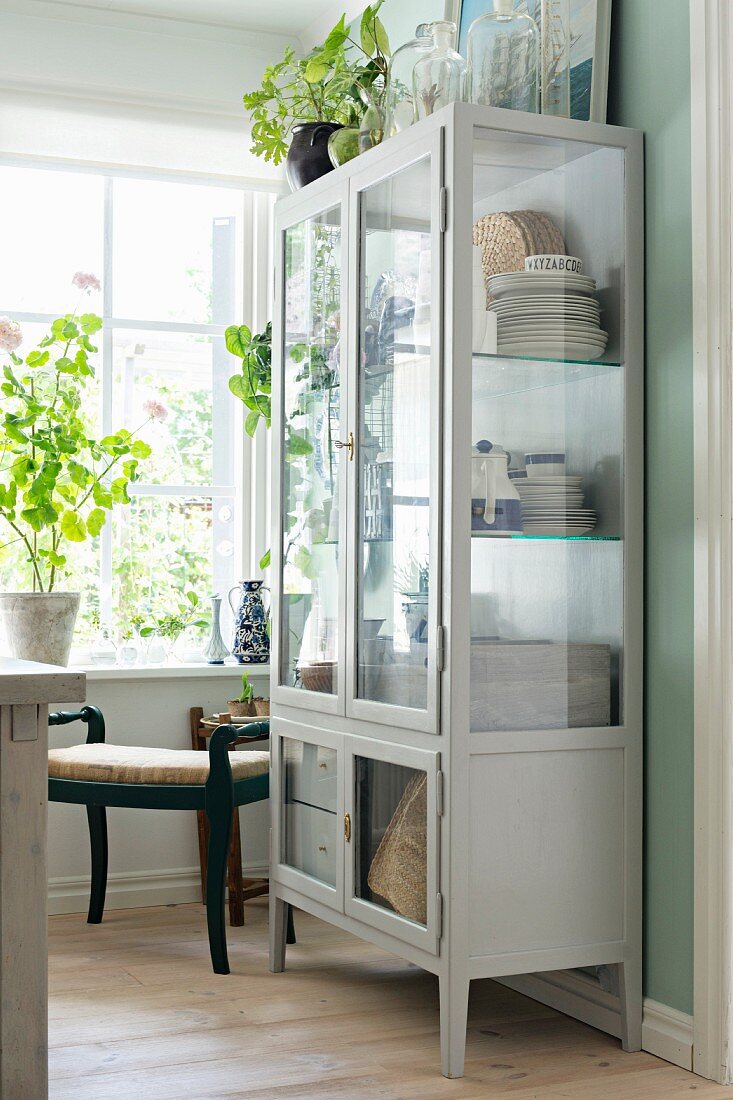 Crockery in white-painted, rustic display case and antique stool in corner next to house plants on windowsill