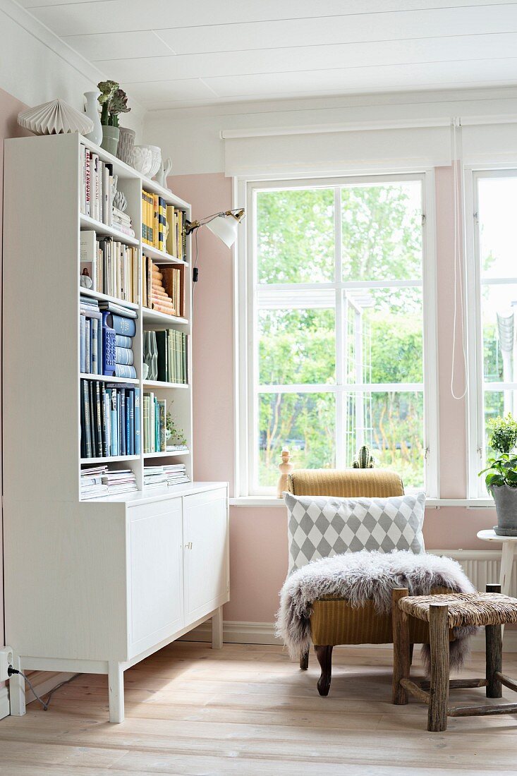 Patterned scatter cushion and fur rug on antique armchair below window and dresser with bookshelves in corner of room painted pink