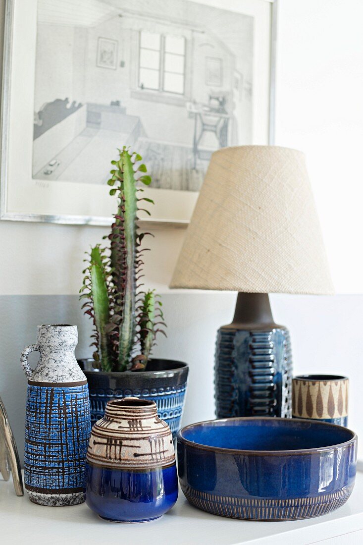 Blaue, gemusterte Keramikbehälter, Kaktus in Übertopf neben Tischleuchte