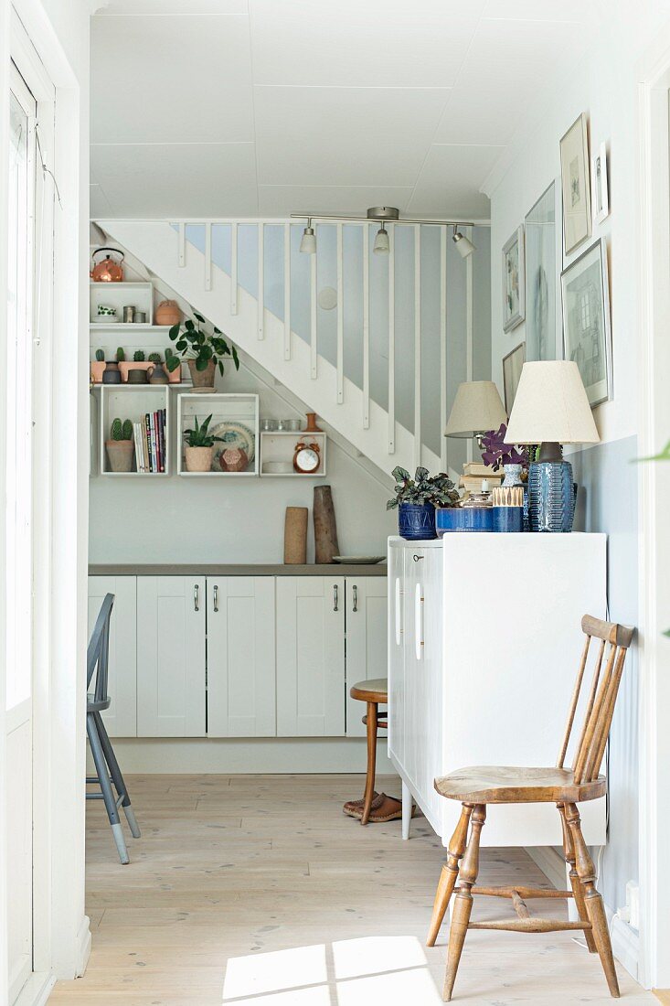 Ländlicher Holzstuhl an Wand neben weisser Kommode in Wohnküche, im Hintergrund Treppenaufgang über Küchenunterschränken