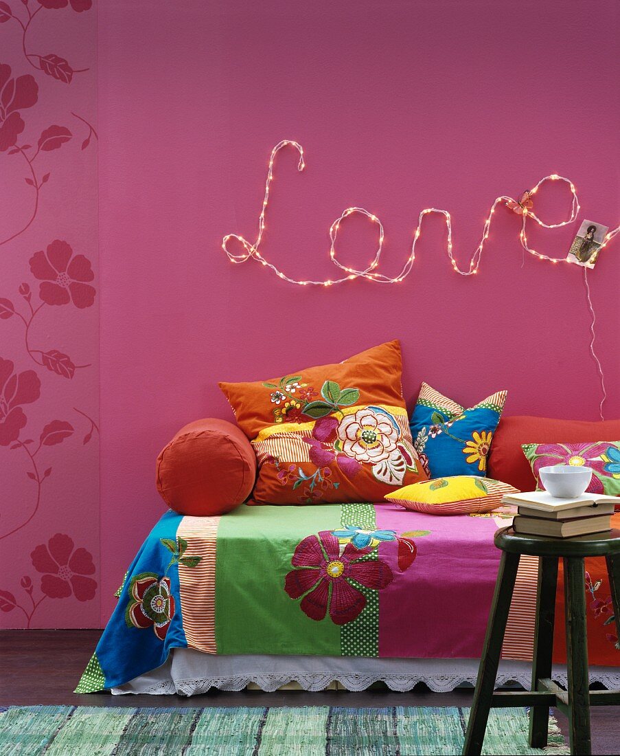 Floral throws and cushions on sofa below motto 'Love' written in fairy lights on deep pink wall