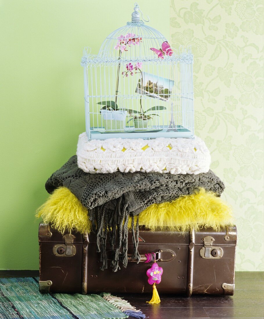 Stacked antique suitcase, cushions, blankets and vintage-style birdcage against pale green wall