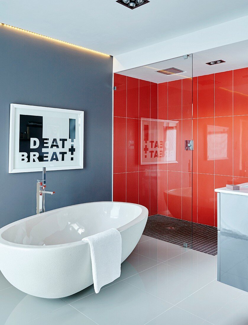 Freistehende weiße Badewanne auf Fliesenboden vor verglastem Duschbereich mit orangeroten Wandfliesen