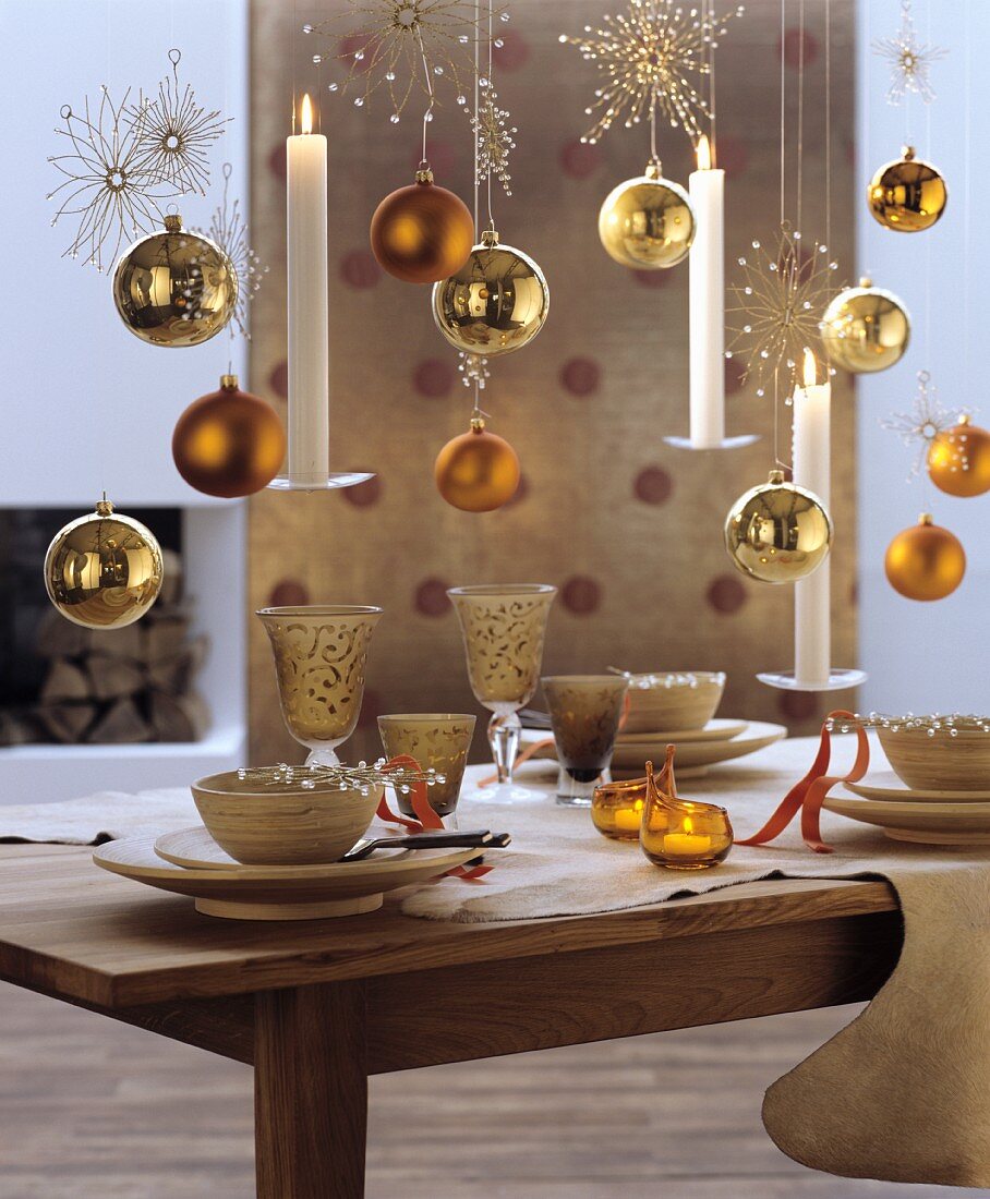 Goldene Baumkugeln, weiße Kerzen und glitzernde Dekosterne aufgehängt über gedecktem Esstisch