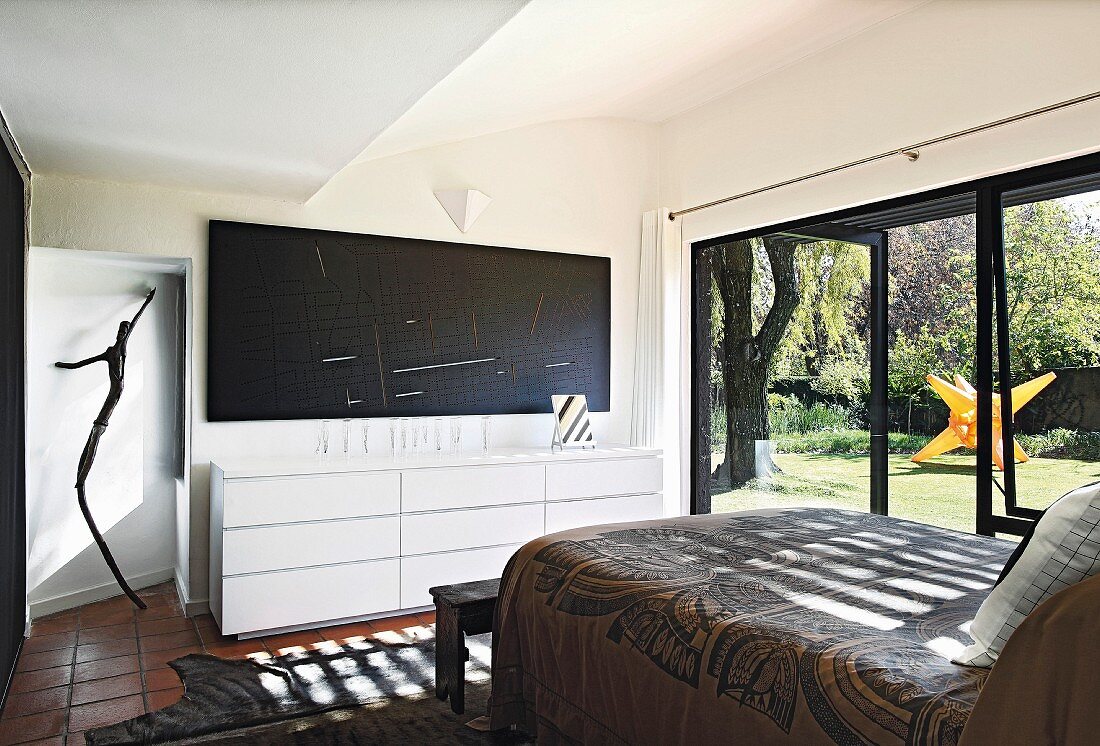 Modern bedroom with box-spring bed opposite sideboard and large TV on wall: terrace doors with view of garden to one side