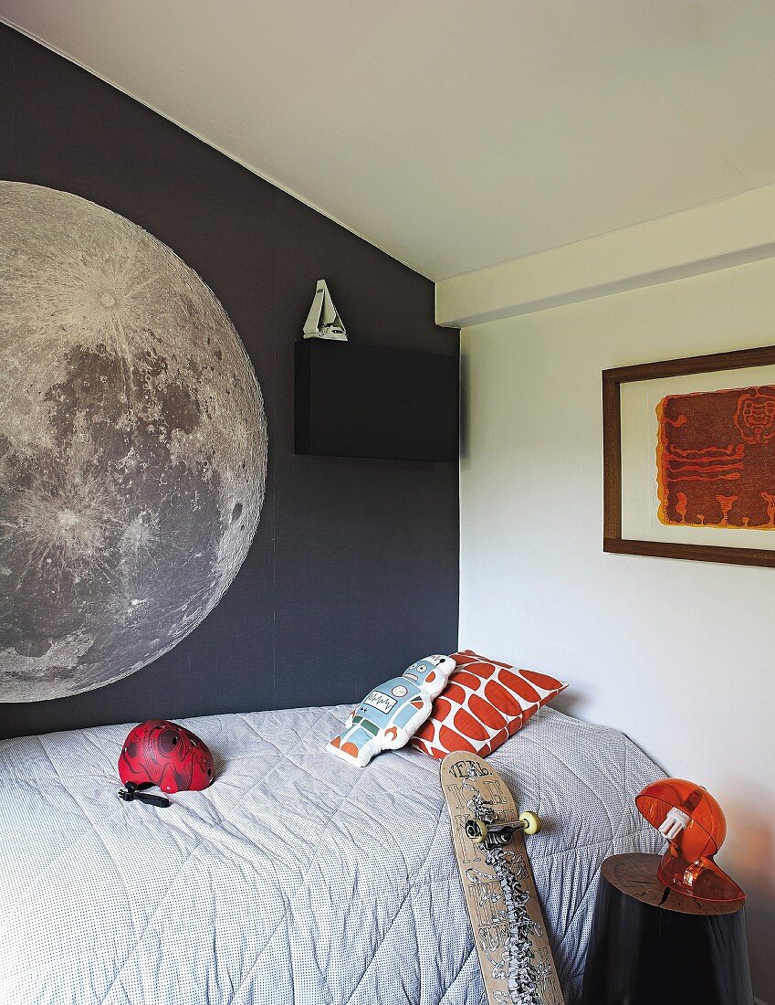 Single bed with pale grey quilt against dark grey wall with mural of moon in teenager's bedroom