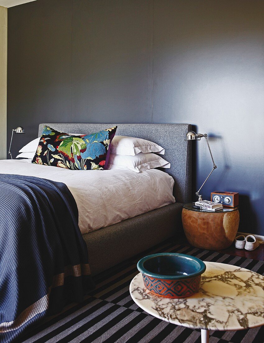 Doppelbett mit buntem Kissen, an schwarz getönter Wand, im Vordergrund Bistrotisch mit Schale