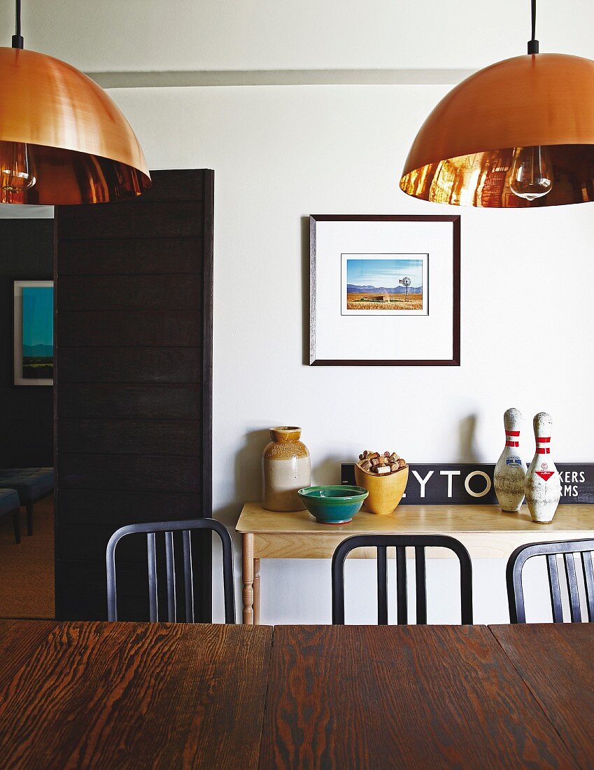 Dark wooden table and chairs below copper pendant lamps in front of various bowls and skittles on console table