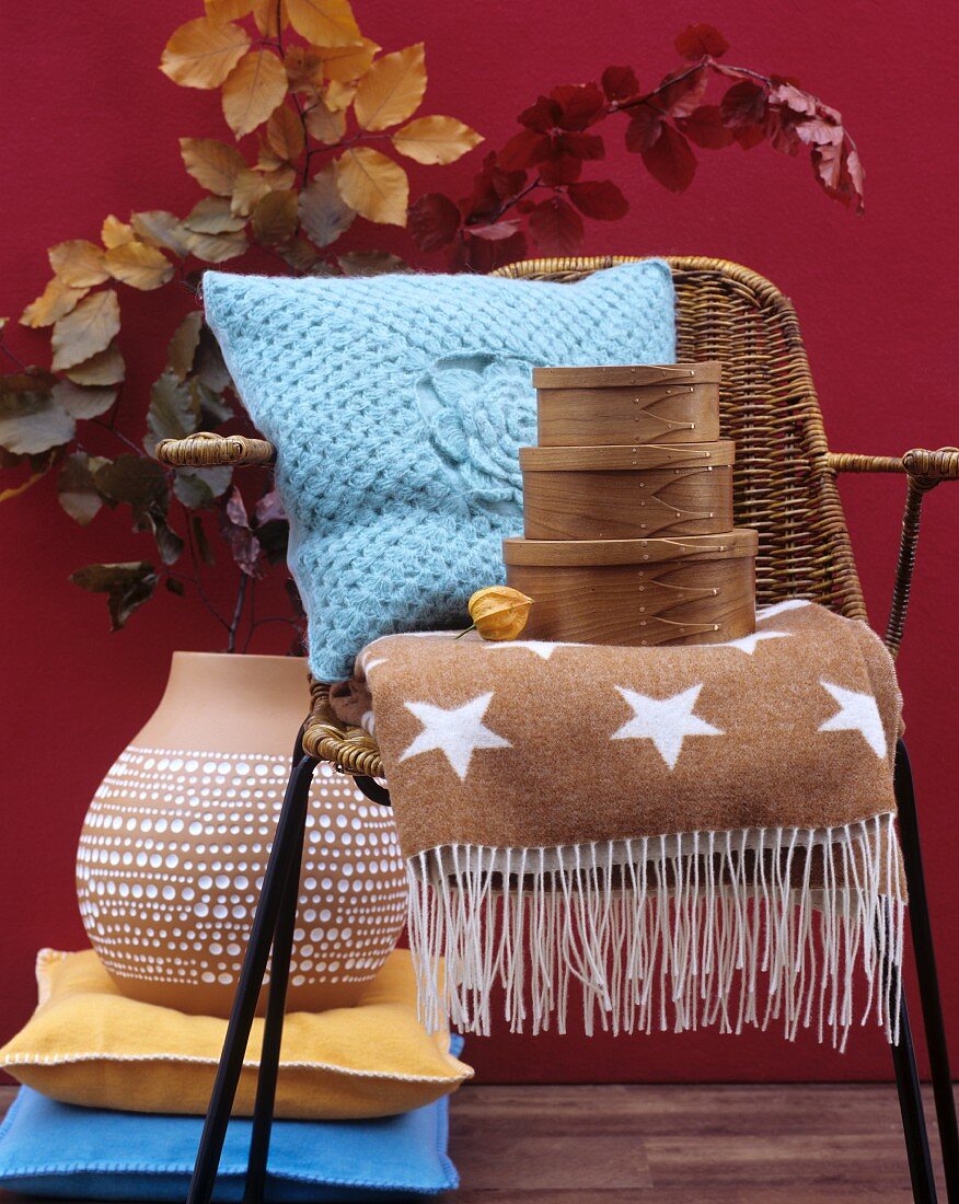 Autumnal home accessories: fifties-style wicker chair, soft cushion, woollen blanket, nest of wooden boxes and terracotta vase