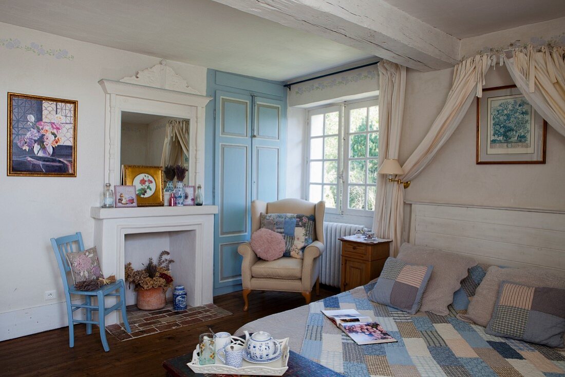 Double bed with patchwork quilt under draped fabric canopy and armchair below window in rustic bedroom