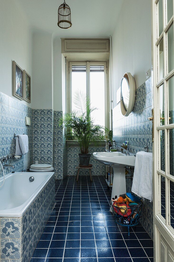 Blick durch offene Tür ins Bad auf dunkelblauen Fliesenboden und gemusterte Fliesen an Wand, im Hintergrund Zimmerpalme auf Schemel vor Fenster