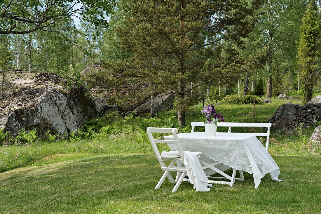 Lace tablecloth on white garden table, bench and chair on lawn with boulders and woods in background