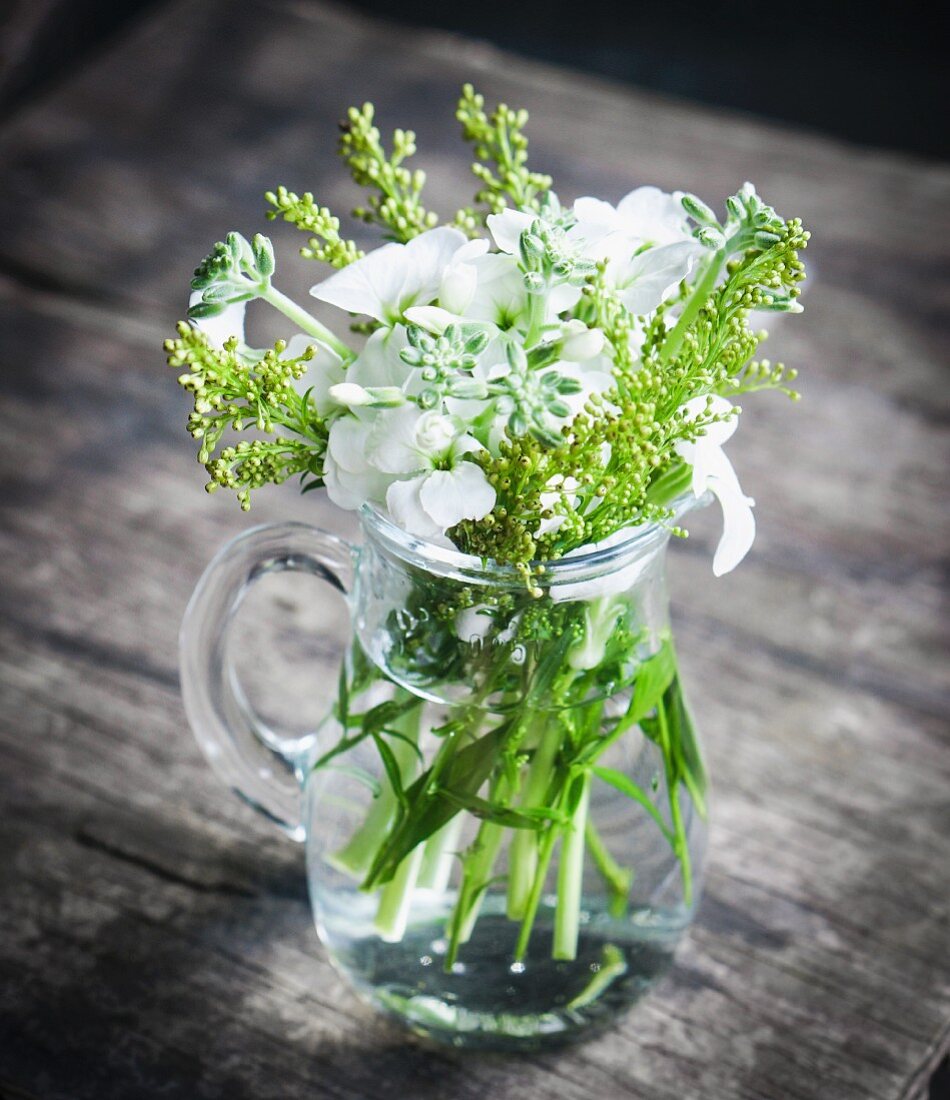 Green stems with buds and white phlox in glass jug on wooden surface