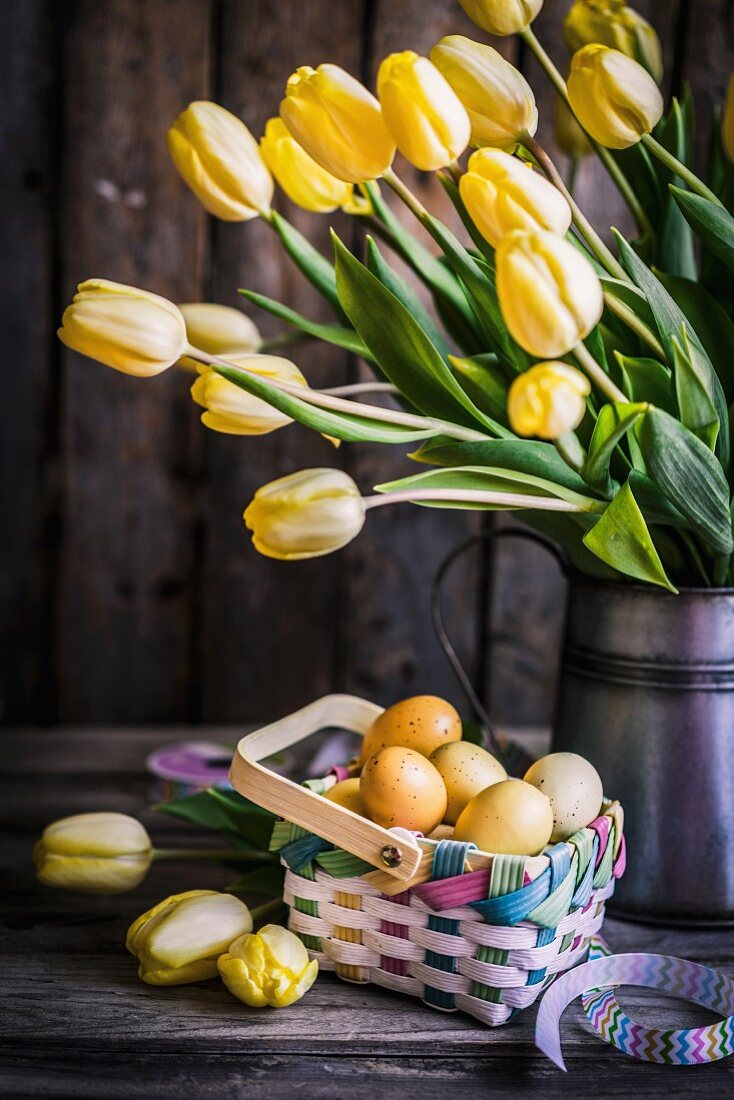 Gelber Tulpenstrauß in Krugvase auf rustikaler Holzunterlage mit pastellfarbenem Flechtkörbchen und Ostereiern