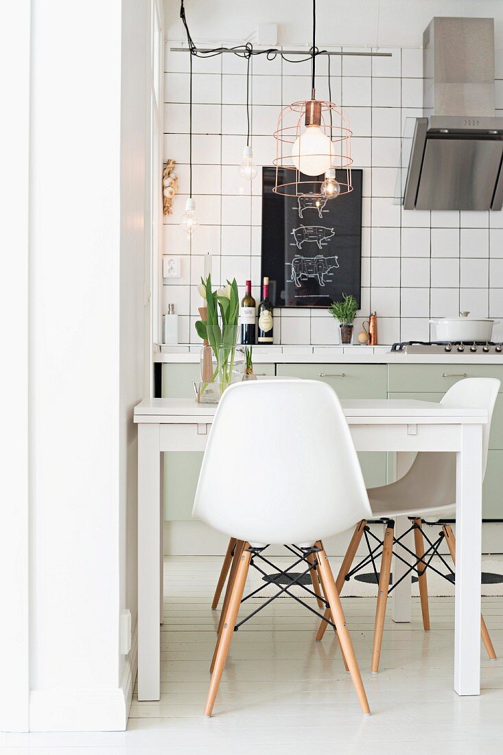 Classic white chairs around modern table below pendant lamp with wire lampshade in kitchen with white-tiled wall