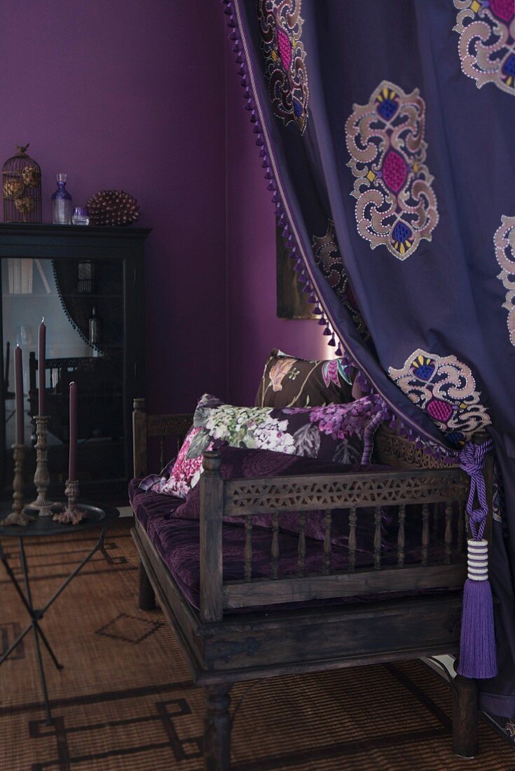 Drapierter Vorhang und antike Sitzbank mit gemusterten Kissen in auberginefarbener Zimmerecke