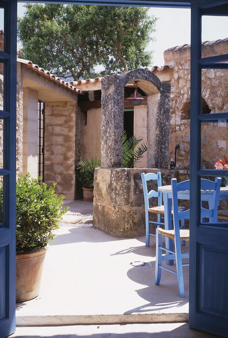 Blick durch offene Terrassentür in sonnenbeschienenen Innenhof, auf Sitzplatz mit blau gestrichenen Stühlen, mediterraner Flair